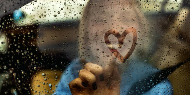 Happy woman drawing heart on car window during rainy day